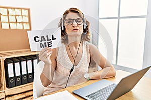 Middle age brunette woman wearing operator headset holding call me banner looking at the camera blowing a kiss on air being lovely