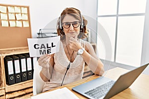 Middle age brunette woman wearing operator headset holding call me banner looking confident at the camera smiling with crossed
