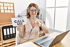 Middle age brunette woman wearing operator headset holding call me banner pointing fingers to camera with happy and funny face