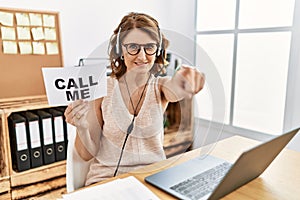 Middle age brunette woman wearing operator headset holding call me banner pointing to you and the camera with fingers, smiling