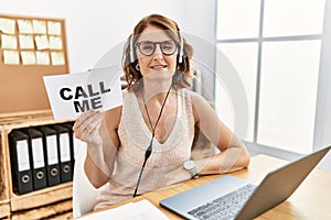 Middle age brunette woman wearing operator headset holding call me banner winking looking at the camera with sexy expression,