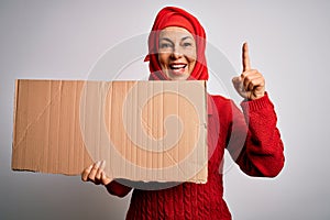 Middle age brunette woman wearing traditional muslim hijab holding banner surprised with an idea or question pointing finger with