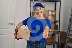 Middle age brunette woman working wearing delivery uniform and cap smiling friendly offering handshake as greeting and welcoming