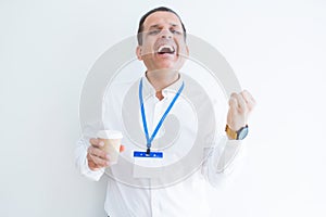 Middle age business man wearing ID card and drinking coffeeover white background screaming proud and celebrating victory and