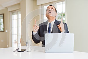 Middle age business man working with computer laptop amazed and surprised looking up and pointing with fingers and raised arms