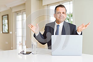 Middle age business man working with computer laptop clueless and confused expression with arms and hands raised