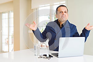 Middle age business man working using laptop clueless and confused expression with arms and hands raised