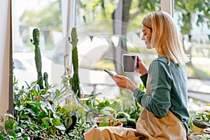 middle age business woman employer sitting with phone near window among flowers, browsing messages or websites in