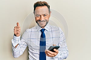 Middle age businessman using smartphone smiling happy and positive, thumb up doing excellent and approval sign