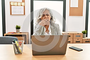 Middle age businesswoman sitting on desk working using laptop at office bored yawning tired covering mouth with hand