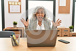 Middle age businesswoman sitting on desk working using laptop at office celebrating crazy and amazed for success with arms raised