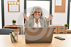Middle age businesswoman sitting on desk working using laptop at office celebrating mad and crazy for success with arms raised and