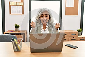 Middle age businesswoman sitting on desk working using laptop at office celebrating surprised and amazed for success with arms