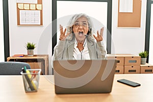 Middle age businesswoman sitting on desk working using laptop at office crazy and mad shouting and yelling with aggressive