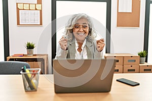 Middle age businesswoman sitting on desk working using laptop at office excited for success with arms raised and eyes closed