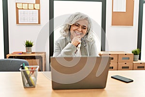 Middle age businesswoman sitting on desk working using laptop at office looking confident at the camera smiling with crossed arms