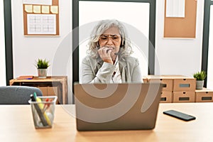 Middle age businesswoman sitting on desk working using laptop at office looking stressed and nervous with hands on mouth biting