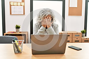 Middle age businesswoman sitting on desk working using laptop at office with sad expression covering face with hands while crying