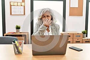 Middle age businesswoman sitting on desk working using laptop at office smelling something stinky and disgusting, intolerable