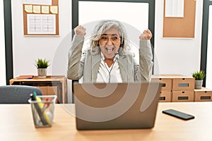 Middle age businesswoman sitting on desk working using laptop at office very happy and excited doing winner gesture with arms