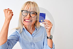 Middle age businesswoman wearing glasses holding credit card over isolated white background annoyed and frustrated shouting with