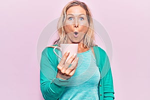 Middle age caucasian blonde woman drinking a cup of coffee scared and amazed with open mouth for surprise, disbelief face