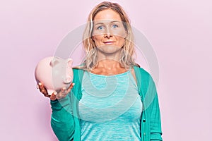 Middle age caucasian blonde woman holding piggy bank thinking attitude and sober expression looking self confident