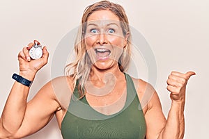Middle age caucasian blonde woman holding stopwatch pointing thumb up to the side smiling happy with open mouth
