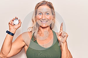 Middle age caucasian blonde woman holding stopwatch smiling with an idea or question pointing finger with happy face, number one