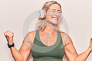 Middle age caucasian blonde woman listening to music using headphones very happy and excited doing winner gesture with arms