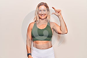 Middle age caucasian blonde woman wearing sportswear smiling and confident gesturing with hand doing small size sign with fingers