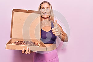 Middle age caucasian blonde woman wearing workout clothes and holding pizza box smiling happy and positive, thumb up doing