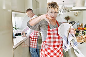 Middle age caucasian couple wearing apron washing dishes at home with angry face, negative sign showing dislike with thumbs down,