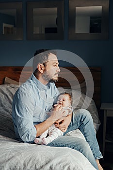 Middle age Caucasian father holding rocking newborn baby on laps. Tired man parent sitting on bed at home with a child on arms