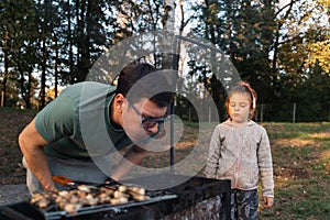 Middle age caucasian father man with child girl blowing air into the bbq coals while cooking grilled champignons