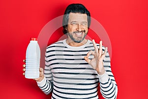 Middle age caucasian man holding liter bottle of milk doing ok sign with fingers, smiling friendly gesturing excellent symbol