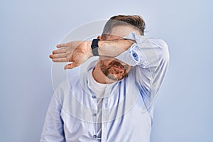 Middle age caucasian man standing over blue background covering eyes with arm, looking serious and sad