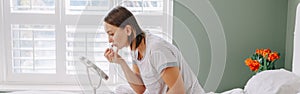 Middle age Caucasian woman applying makeup cosmetics in morning. Young woman sitting in bed at home bedroom doing her morning