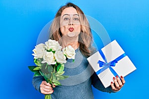 Middle age caucasian woman holding anniversary present and bouquet of flowers looking at the camera blowing a kiss being lovely