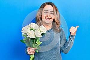 Middle age caucasian woman holding flowers pointing thumb up to the side smiling happy with open mouth