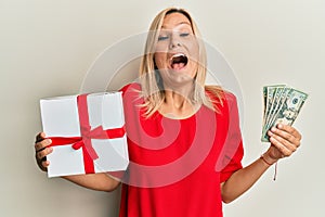 Middle age caucasian woman holding gift and dollars celebrating crazy and amazed for success with open eyes screaming excited