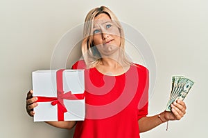 Middle age caucasian woman holding gift and dollars in shock face, looking skeptical and sarcastic, surprised with open mouth