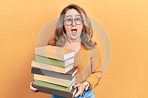 Middle age caucasian woman holding a pile of books afraid and shocked with surprise and amazed expression, fear and excited face