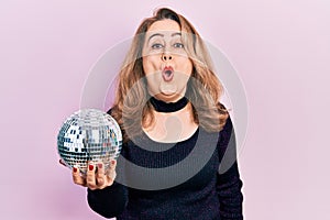 Middle age caucasian woman holding shiny disco ball scared and amazed with open mouth for surprise, disbelief face