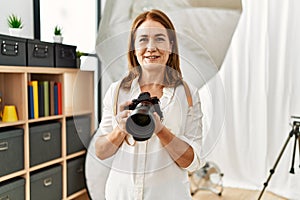 Middle age caucasian woman photographer smiling confident holding professional camera at photograph studio