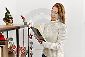 Middle age caucasian woman reading book standing by christmas decor at home