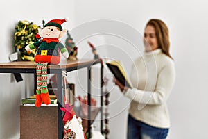 Middle age caucasian woman reading book standing by christmas decor at home