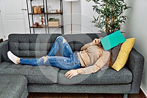 Middle age caucasian woman sleeping on the sofa covering face with book at home