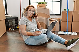 Middle age caucasian woman using laptop and talking on the smartphone sitting on the floor at new home