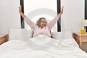 Middle age caucasian woman waking up stretching arms sitting on the bed at bedroom